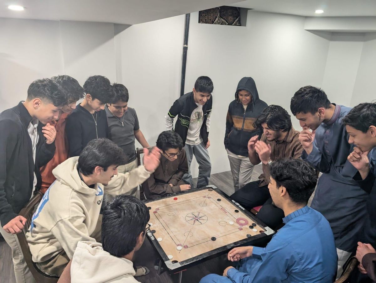 A dozen people crowded around a carrom board in a room, mid game.
