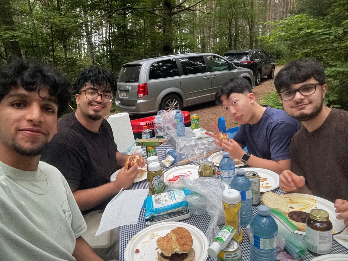 Five of us around a folding table of food at a wooded campsite, cars parked behind.