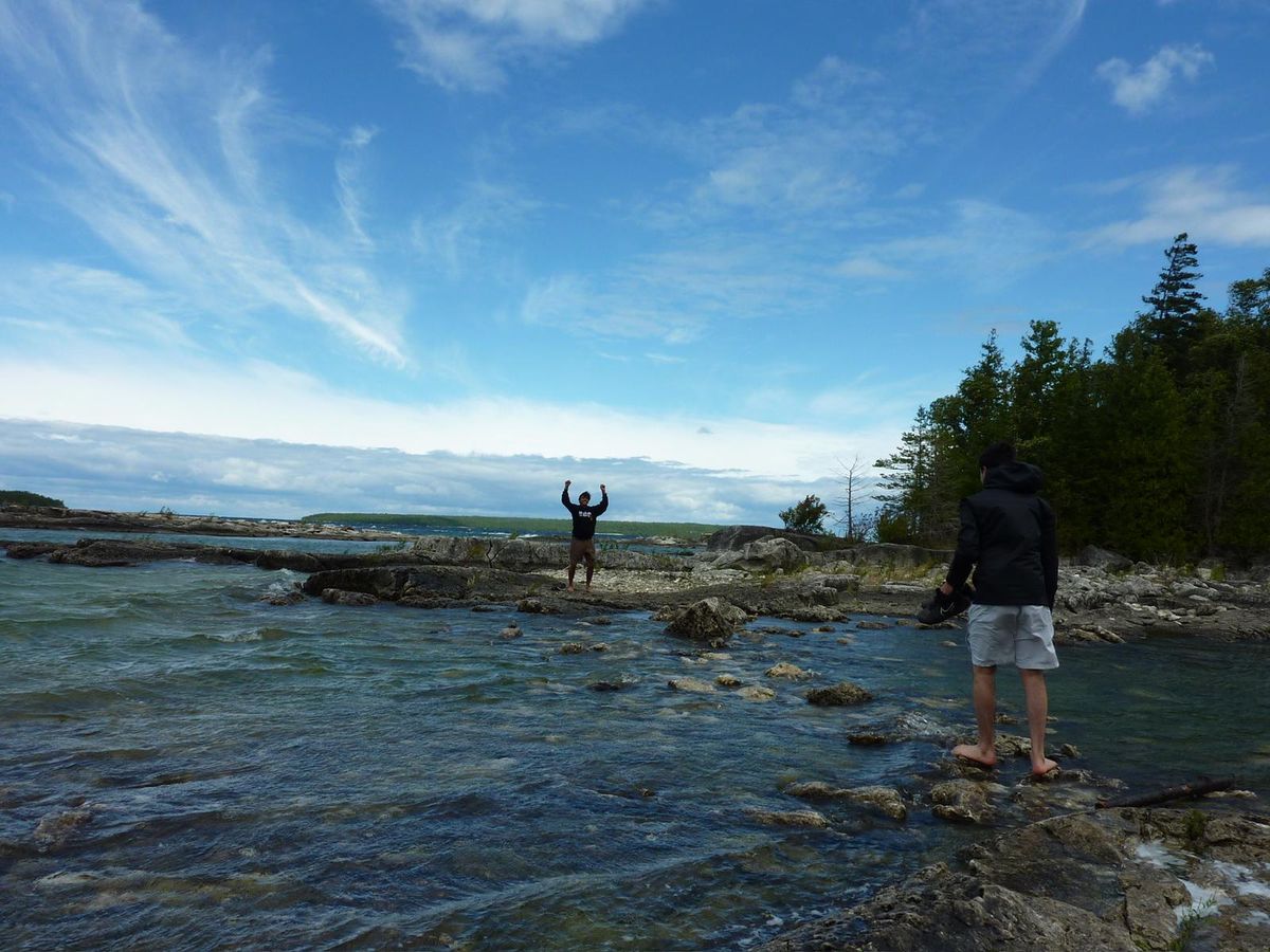 Two people on a rocky shoreline beside clear shallow water under a wide sky.
