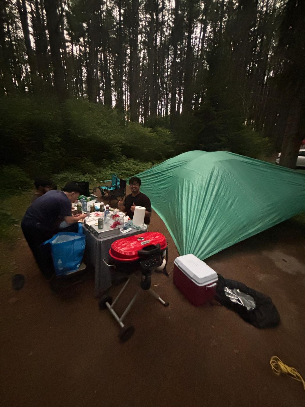 A campsite at dusk: a green tent, a folding table with a grill and a cooler, friends sitting around it.