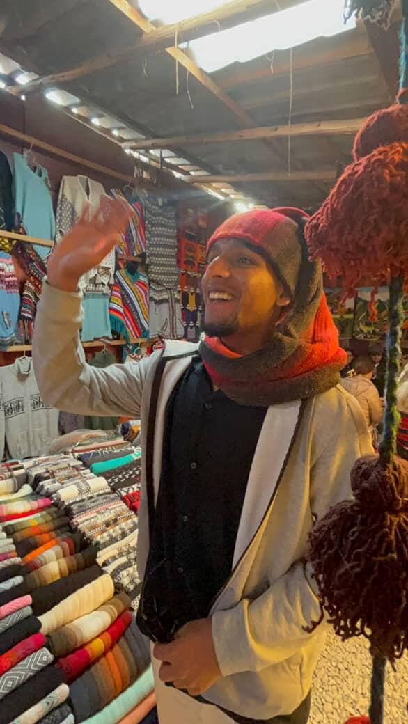 In a market stall hung with woven blankets and bundles of dyed wool, wearing a knitted hat and scarf.