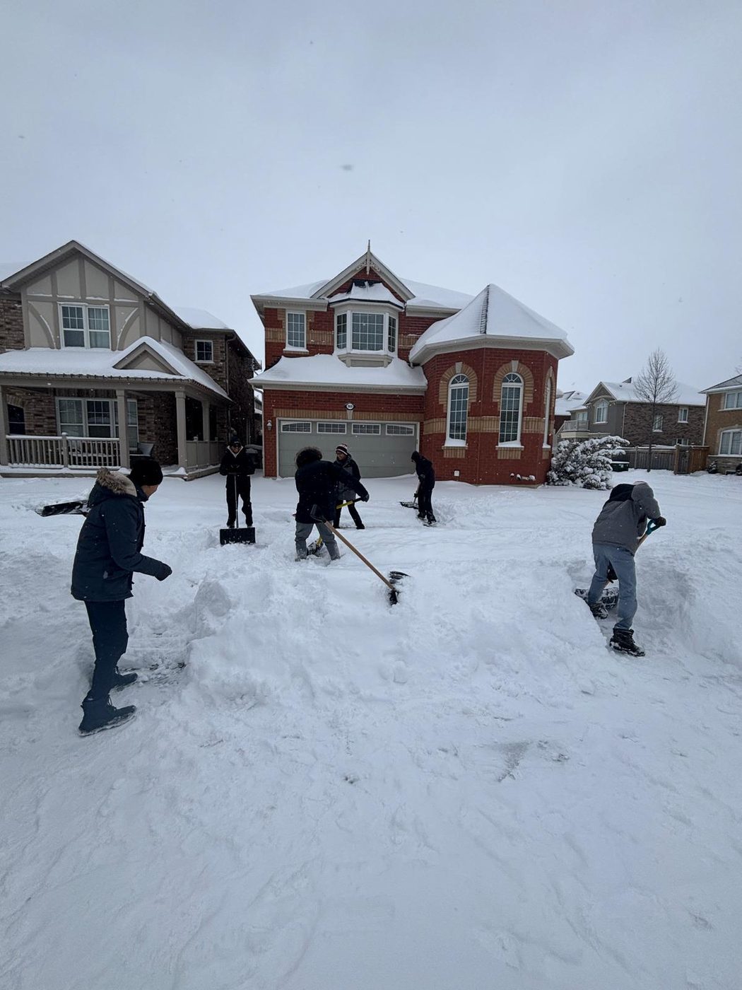 Six people with shovels clearing a driveway and front lawn of a brick house buried in deep snow.