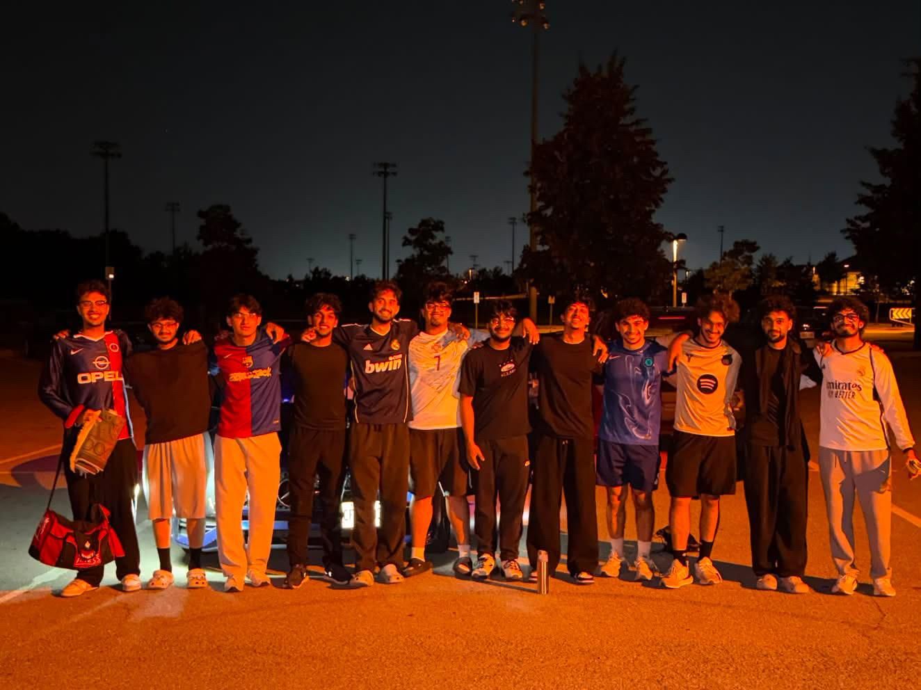 A dozen players in assorted club shirts lined up arm in arm on a floodlit parking lot after a night game.