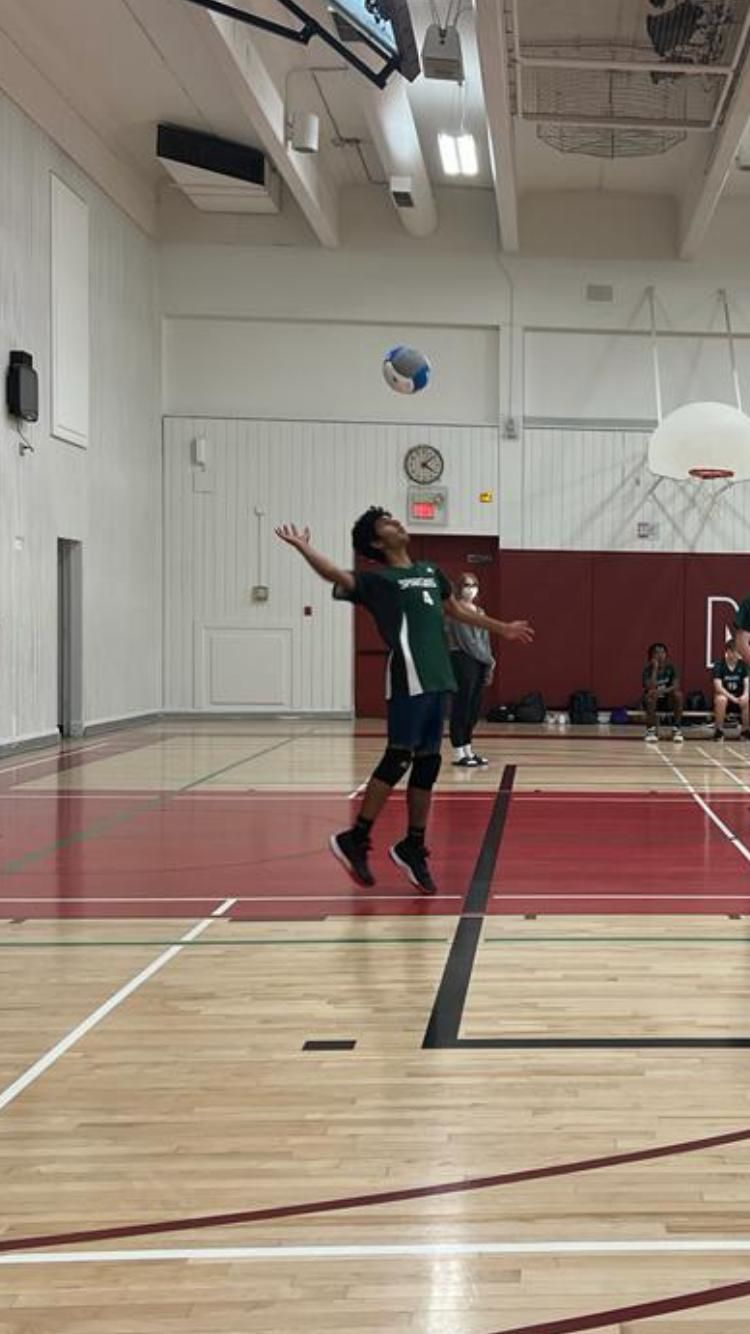 Mid jump serve in a school gym, ball tossed overhead, green jersey and knee pads.
