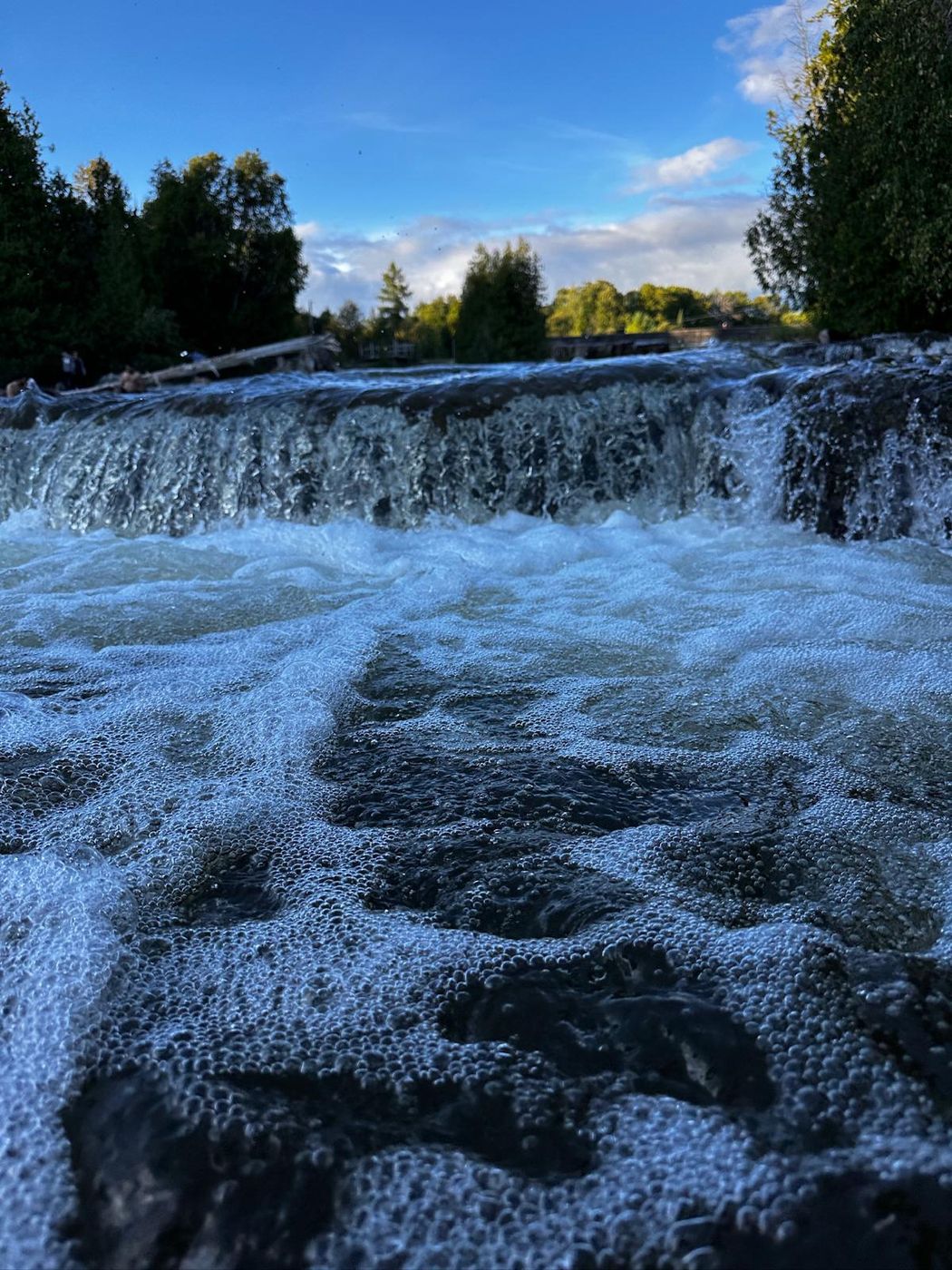 A wide low waterfall spilling over a rock shelf into fast white water, trees and blue sky above.